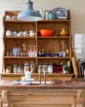 Woman preparing food in a rustic kitchen with wooden shelves, dishes, and a large orange pot.