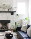 Living room with white stone fireplace, black sofa, armchair, plants, and a wooden coffee table.