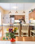 Open kitchen with wooden cabinets, island with bar stools, pendant lights, and a person pouring a drink.