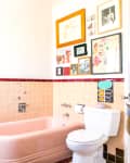 Vintage bathroom with pink bathtub, red-tiled trim, eclectic wall art, wooden chair with red cushion, and white toilet.