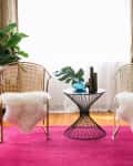 Two wicker chairs with white fur throws, black wire table, plants, and a guitar on a pink rug in a sunlit room.