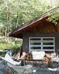 Cabin exterior with large glass garage doors, surrounded by trees, firewood stack, and white butterfly chairs on a patio.