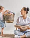 Child handing a basket to a woman sitting on the floor, both smiling, with folded clothes and a cozy rug.