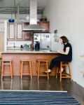 Modern kitchen with wooden island, four stools, stainless steel appliances, and a person working on a laptop.