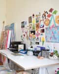 Art studio with colorful paintings, two wooden stools, a white desk, printer, and vibrant wall art display.