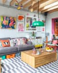 Colorful living room with gray sofa, vibrant cushions, striped rug, wooden coffee table, and eclectic wall art.