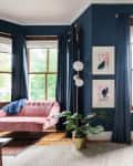 Living room with pink sofa, blue walls, large mirror, potted plant, and abstract art near a window with blue curtains.