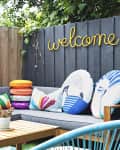Outdoor patio with gray sectional, colorful pillows, wooden table, and "welcome" sign on fence. Two vibrant wall art pieces.