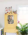 Bright room with yellow cabinet, potted plants, framed art, and a white shelf with a lamp and decor items.