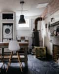 Industrial-style kitchen with a wooden island, white chairs, hanging lights, and a vintage stove next to a fireplace.