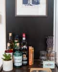 Home bar setup with assorted liquor bottles, cocktail books, a potted plant, and lavender in jars under a framed tree artwork.