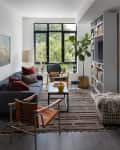 Modern living room with a blue sofa, striped rug, bookshelves, and large window overlooking greenery.