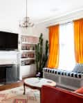 Living room with orange curtains, cactus, neon heart light, bookshelves, and a wall-mounted TV above a fireplace.
