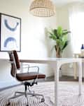 Home office with a brown swivel chair, white desk, large plant, and abstract art on the wall.