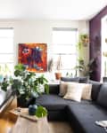 Studio apartment with a bed, dark sectional sofa, vibrant abstract painting, and various potted plants by the windows.