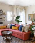 Cozy living room with a coral sofa, colorful cushions, plants, and a round dining table with art on the walls.