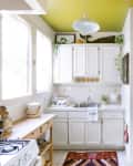 Bright kitchen with white cabinets, yellow ceiling, colorful rug, and plants on countertops and shelves.