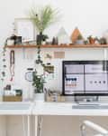 Home office with white desk, computer, plants, art, and decor on shelves, creating a creative and organized workspace.