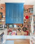 Kitchen with exposed brick, blue cabinets, marble countertop, hanging pots, and a fridge covered in magnets and notes.