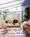 Sunroom with yellow sofa, colorful cushions, wicker chair, wooden cabinet, and patterned rug under a glass roof.