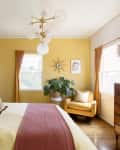 Cozy bedroom with mustard yellow walls, a bed with neutral pillows, a mustard armchair, and a modern chandelier.