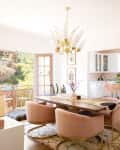 Dining room with pink chairs, wooden table, gold chandelier, macramé wall hanging, and open French doors to a garden.