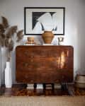 Vintage wooden dresser with abstract art, wicker vase, books, and pampas grass in a white vase on a wooden floor.