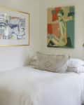 Cozy bedroom with abstract art, white bedding, gold floor lamp, and potted plant by a window.