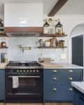 Modern kitchen with blue cabinets, marble countertops, open shelves, brass fixtures, and a black stove under a white hood.