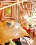 Bedroom with geometric red and yellow wall patterns, a bed with a skull pillow, and a red side table with a lamp.