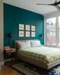 Bedroom with teal accent wall, framed art, green patterned bedspread, large windows, and a blue patterned rug.
