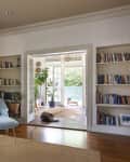 Living room with blue chair, bookshelves, hanging plant, and open doors to sunroom with potted plants and a dog.