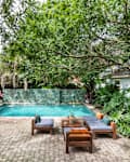 Backyard with a turquoise pool, surrounded by lush greenery, brick patio, and wooden outdoor furniture with cushions.