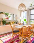 Dining room with round table, wicker chairs, colorful rug, plants, and large windows with sheer curtains.