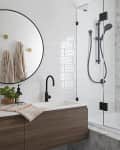 Modern bathroom with round mirror, dual sinks, black faucets, wood vanity, and glass shower with hexagonal tile wall.