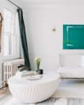 Bright living room with white sofa, teal chair, round coffee table, and abstract green painting above.
