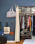 Open closet with assorted clothes, bags, and shoes in a blue room, next to a red sofa with patterned pillows.