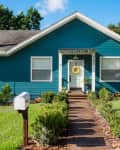 Teal house with white trim, wooden walkway, mailbox, and yellow wreath on door, surrounded by green lawn and shrubs.