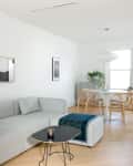 Modern living room with a light gray sofa, black floor lamp, abstract wall art, and a round dining table with wooden chairs.