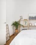 A white bedroom with a wood bed frame and a wooden shelf along the wall, plus a small stool with a plant on it in the corner.