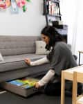 Woman kneeling, organizing books in a drawer under a gray sofa in a living room with wall art and shelves.