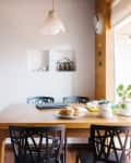 Dining area with wooden table, black chairs, pendant light, muffins, and potted plants by a window.