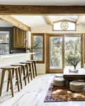 Open living room with wooden beams, bar stools, large windows, a low table, floor cushions, and a colorful rug.
