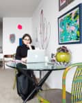 Woman working on a laptop at a glass table in a modern living room with colorful artwork and a potted plant.
