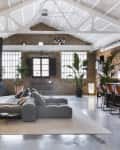 Spacious loft with exposed brick walls, large windows, gray sectional sofa, and indoor plants.