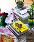 Glass coffee table with stacked books, a small plant in a green vase, colorful rug, and a black iridescent cup.
