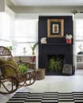 Cozy living room with a black fireplace, wicker chair, leather sofa, plants, and a striped rug.