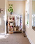 Clothing rack with white shirts and floral dress, shelves with bags, baskets, and a potted plant in a bright room.