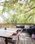Wooden deck with a large table, pink chairs, string lights, potted plants, and a black outdoor sofa.