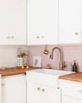 White kitchen with pink subway tile backsplash, brass faucet, mint kettle, and citrus-patterned towel on stove.